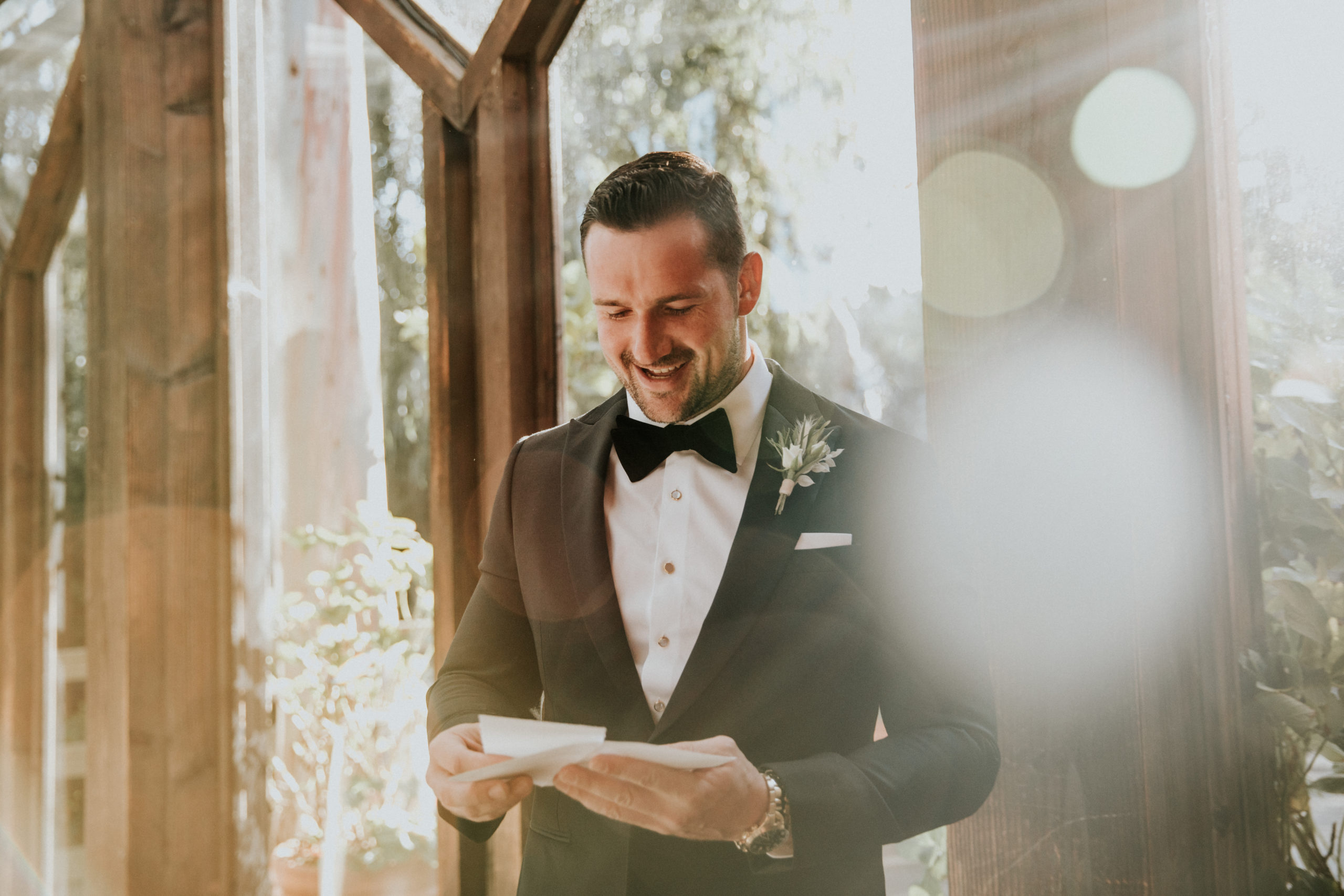 Groom getting ready on wedding day with light beaming through the window reading a letter from his bride smiling