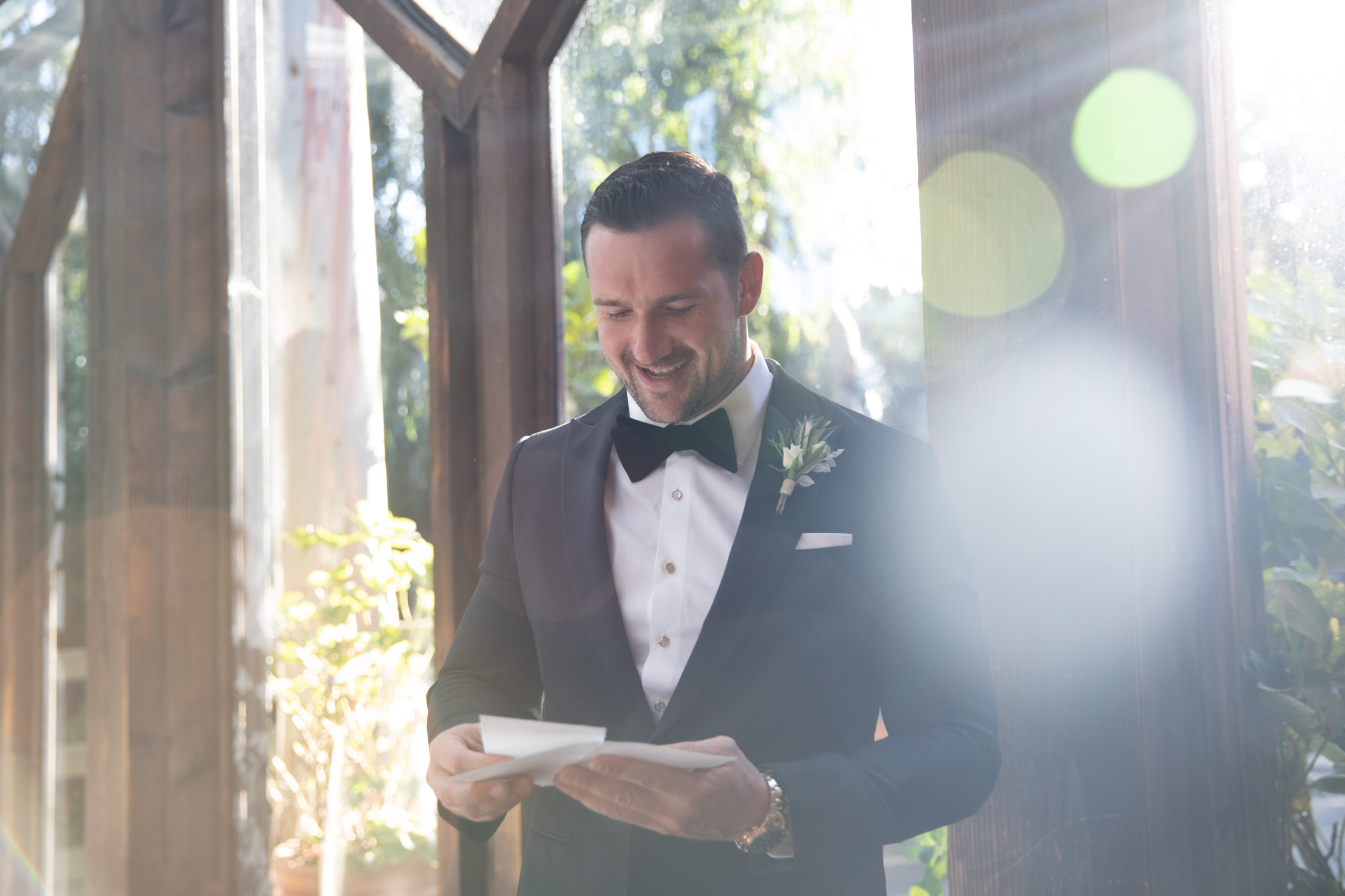 Groom getting ready on wedding day with light beaming through the window reading a letter from his bride smiling