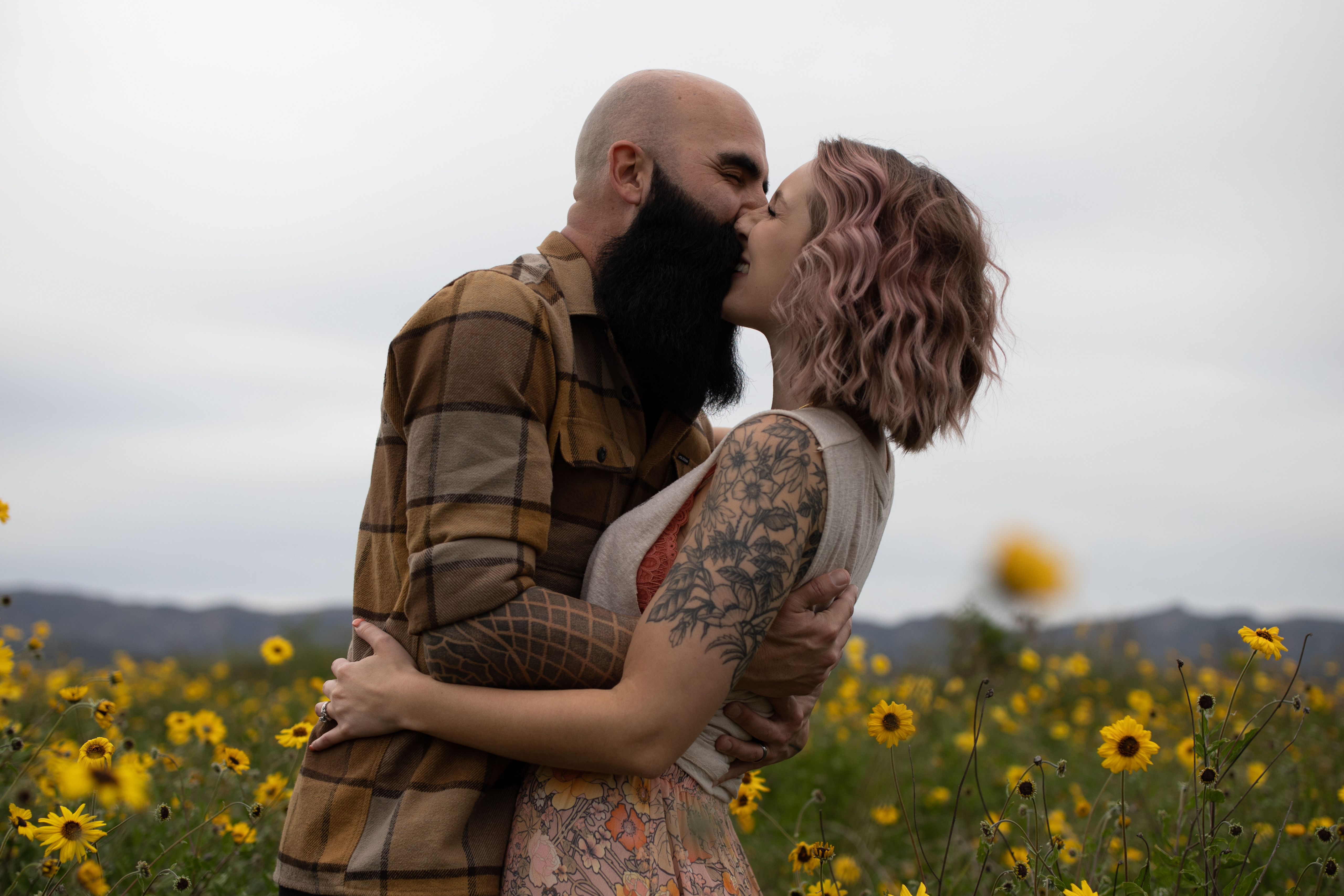Husband and wife couple laughing and kissing with tattoos and beard and pink hair in yellow daisy field